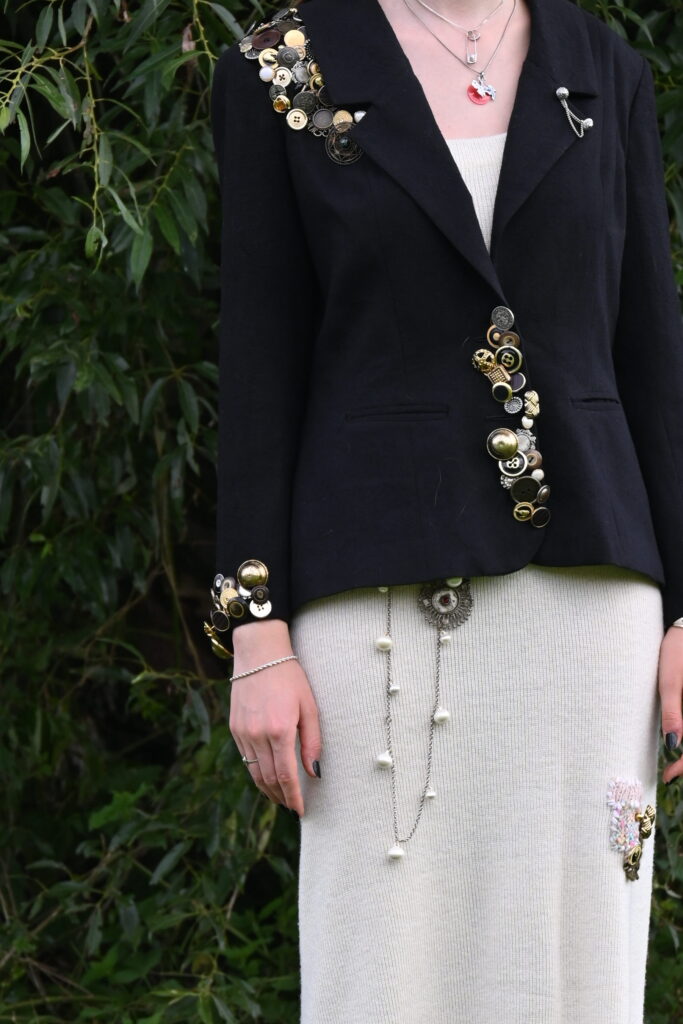 Reichlich verzierter schwarzer Blazer mit vielen goldenen Knöpfen und weißem Strickkleid mit weiteren Verzierungen